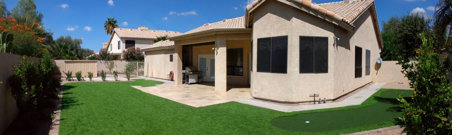 Outdoor view of a single-story assisted living facility building with a tiled roof, surrounded by a well-maintained lawn and a small putting green. The area includes a covered patio with outdoor furniture and is enclosed by a fence and greenery under a clear blue sky.