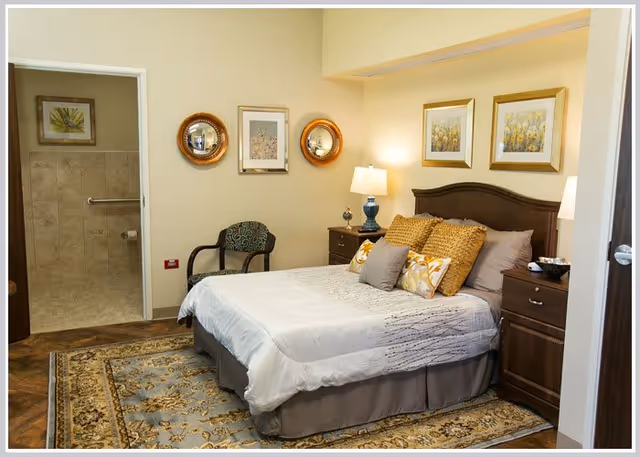 A cozy bedroom with a neatly made bed featuring multiple pillows and a patterned bedspread. The room has two wooden nightstands with lamps on each side of the bed. There is a patterned armchair against the wall, which is decorated with framed artwork and two round mirrors. An open doorway leads to a bathroom with tiled walls and a grab bar next to the toilet.
