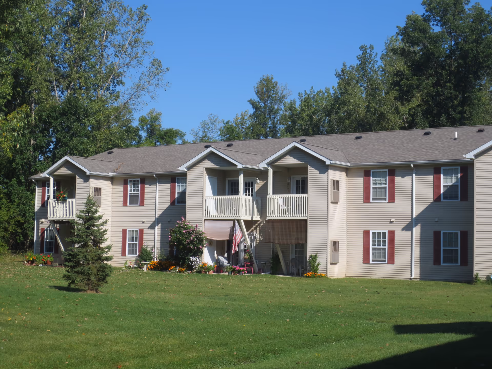 Exterior view of a two-story beige apartment building with red shutters and small balconies, surrounded by green grass and trees under a clear blue sky.