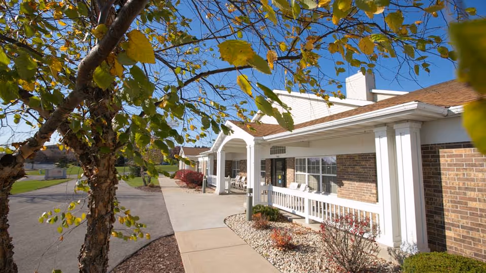 Exterior view of Pleasant Point facility showing a brick building with white columns and a covered porch. There are shrubs and small plants along the walkway, and a tree with green and yellow leaves in the foreground under a clear blue sky.