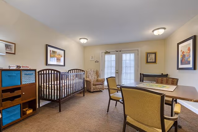 A well-lit room with a crib, an armchair, a table with four chairs, and framed artwork on the walls. The room has beige walls and carpeted flooring, with double glass doors letting in natural light.