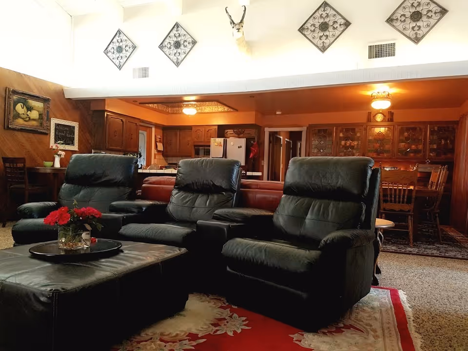A cozy living room area with three black leather recliners arranged around a large black ottoman with a tray holding a vase of red flowers. The room features wood-paneled walls, decorative wall art, and a mounted deer head. In the background, there is a kitchen with wooden cabinets and a dining area with a wooden table and chairs.