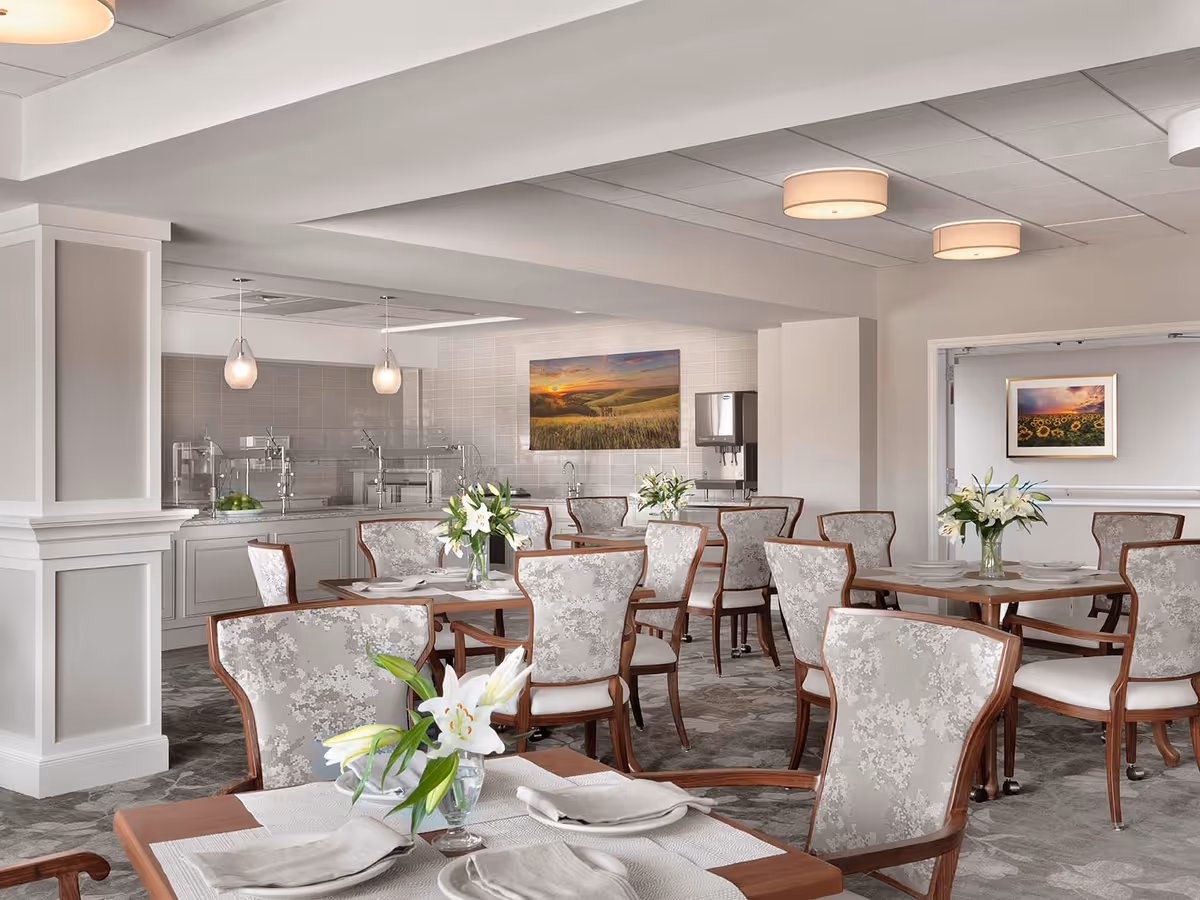 A bright and elegant dining room with several wooden tables and upholstered chairs arranged neatly. Each table is set with white napkins and plates, and decorated with vases of white lilies. The room features soft lighting with ceiling fixtures and pendant lights over a buffet area in the background. The walls are light-colored with framed landscape artwork, and the floor is carpeted in a subtle pattern.
