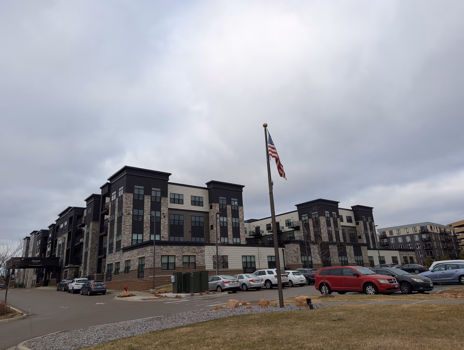 Exterior view of a multi-story senior living facility named Amira Choice Bloomington with a parking lot in front and an American flag on a flagpole. The building has a modern design with stone and dark panel accents under a cloudy sky.