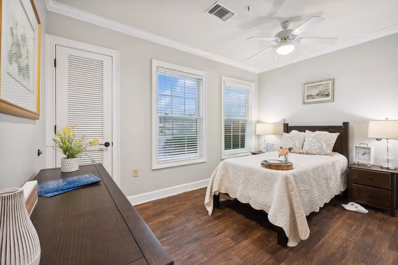 A bright bedroom with a wooden bed frame and a white quilted bedspread. The bed has two pillows and a tray with a small flower arrangement on it. There are two windows with white blinds letting in natural light. On either side of the bed are dark wooden nightstands with lamps and decorative items. A ceiling fan with a light is mounted on the white ceiling. A wooden dresser with a vase of yellow flowers and framed artwork is visible on the left side of the room. The floor is dark wood, and the walls are painted light gray.