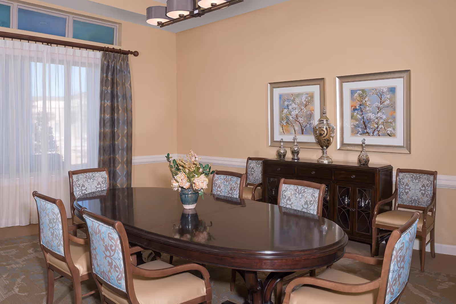A formal dining room with a dark wooden oval table surrounded by eight upholstered chairs with patterned backs. A vase with flowers is centered on the table. Behind the table is a dark wooden sideboard with decorative vases and two framed floral paintings hanging above it. The room has beige walls, a window with sheer white curtains and patterned drapes, and a modern chandelier overhead.