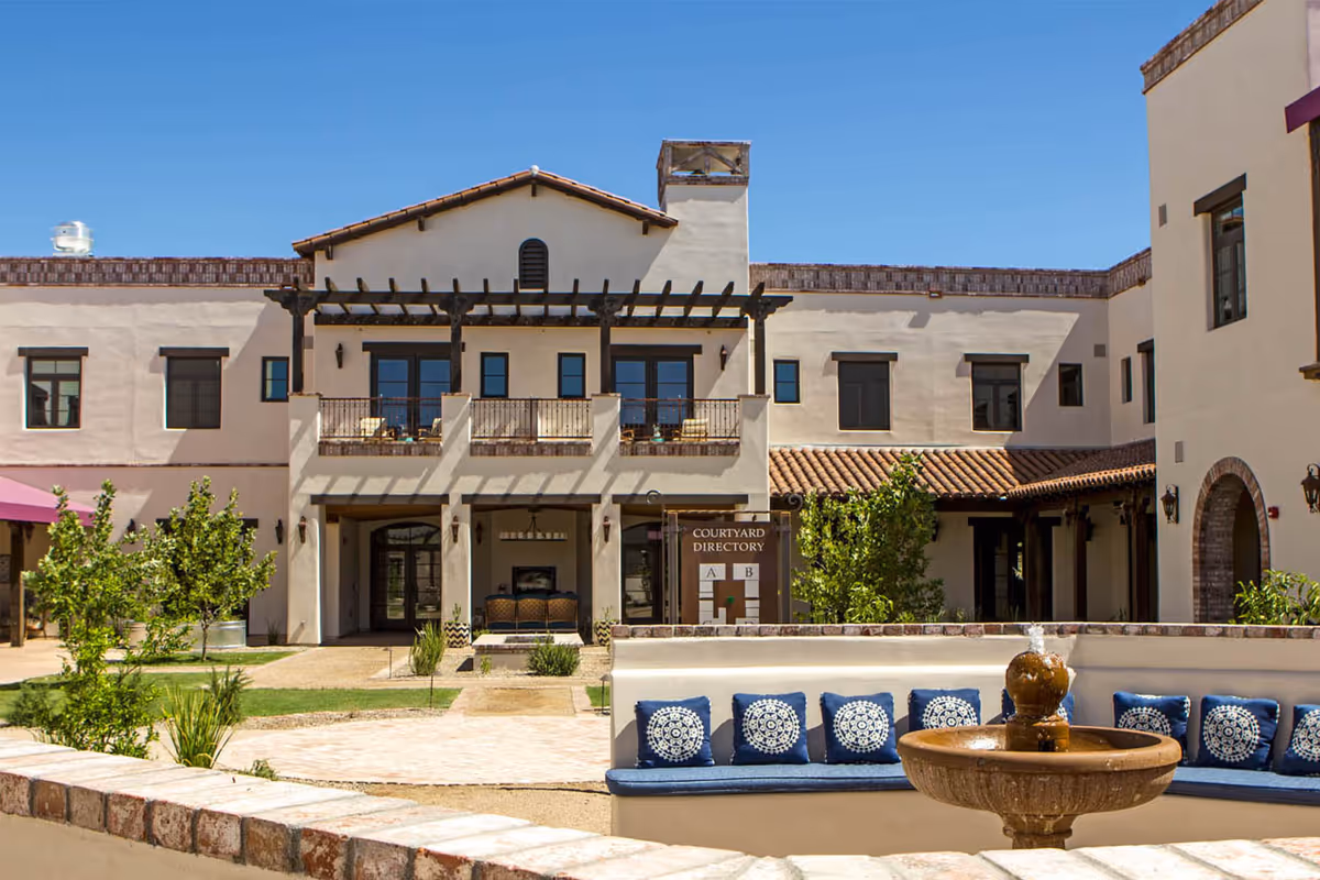 Courtyard with a fountain, blue cushioned seating, and landscaping in front of a two-story Mediterranean-style senior living building.