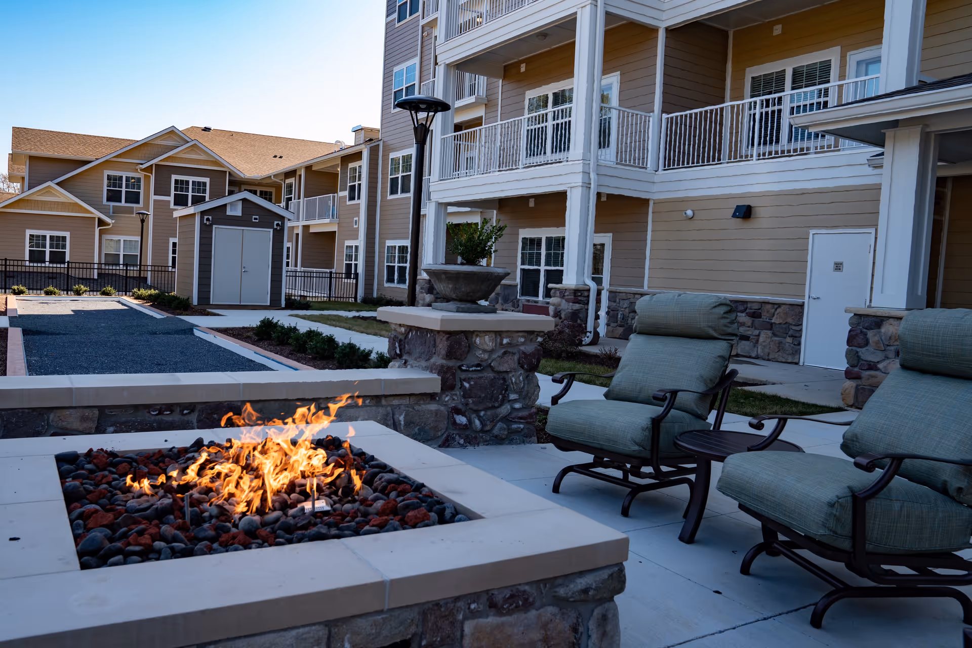 Outdoor courtyard of a senior living building with a lit stone fire pit, cushioned patio chairs, and apartment balconies.