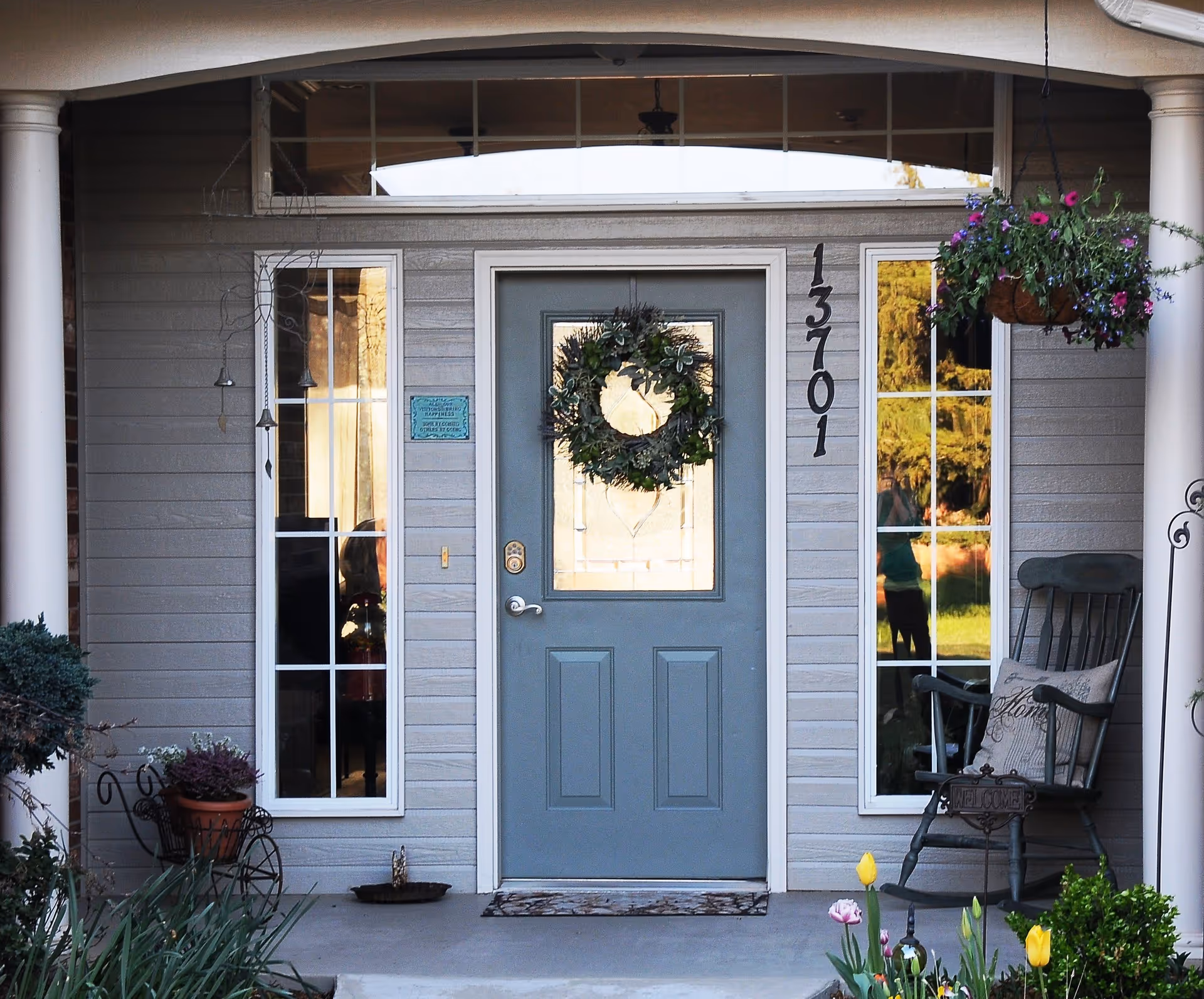Front entrance of a house with a blue door decorated with a green wreath. The house number 13701 is displayed vertically to the right of the door. There are two tall windows on either side of the door, a hanging flower basket with purple and pink flowers on the right, a rocking chair with a cushion, and various potted plants and flowers near the entrance.
