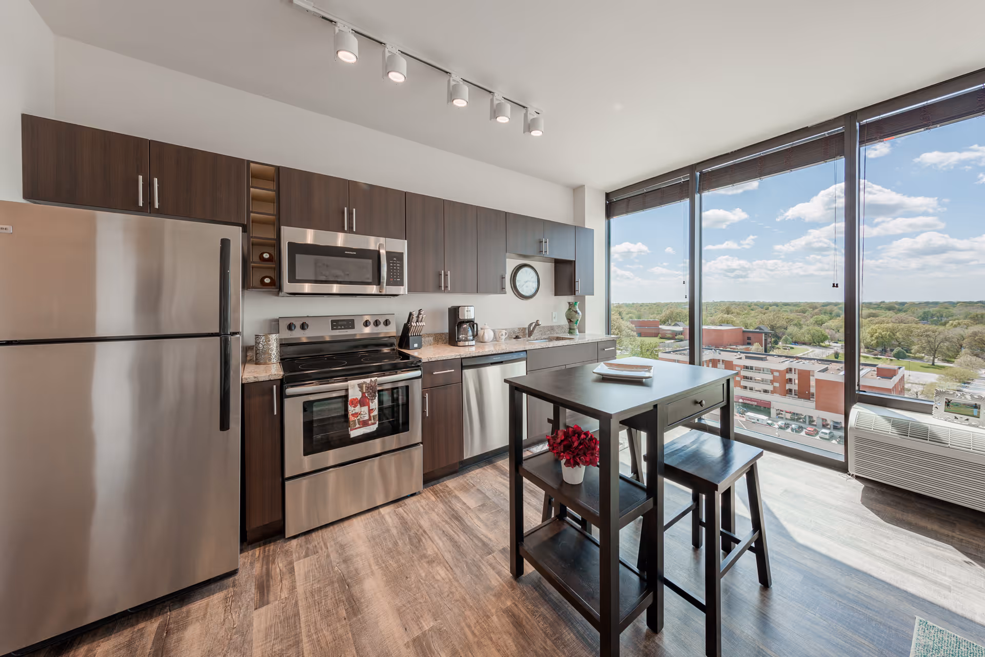 Modern kitchen with stainless steel refrigerator, oven and microwave, dark cabinets, a small dining island and large floor-to-ceiling windows overlooking city buildings and trees.