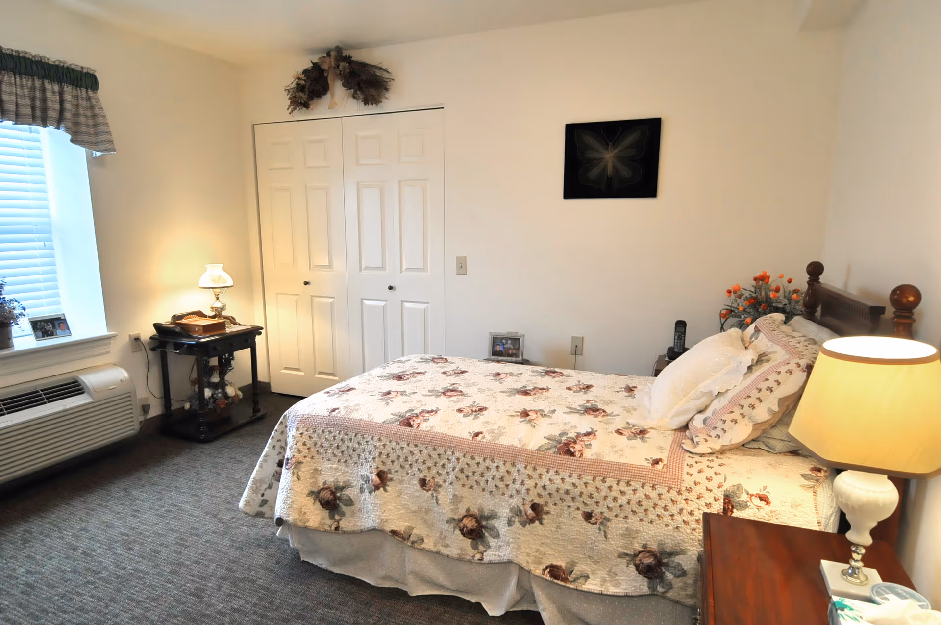 A cozy bedroom with a floral quilted bedspread on a wooden bed frame, a wooden nightstand with a lamp, a small table with a lamp and decorative items, a window with blinds and a valance, and a closed white closet door. The walls are plain white with minimal decoration.
