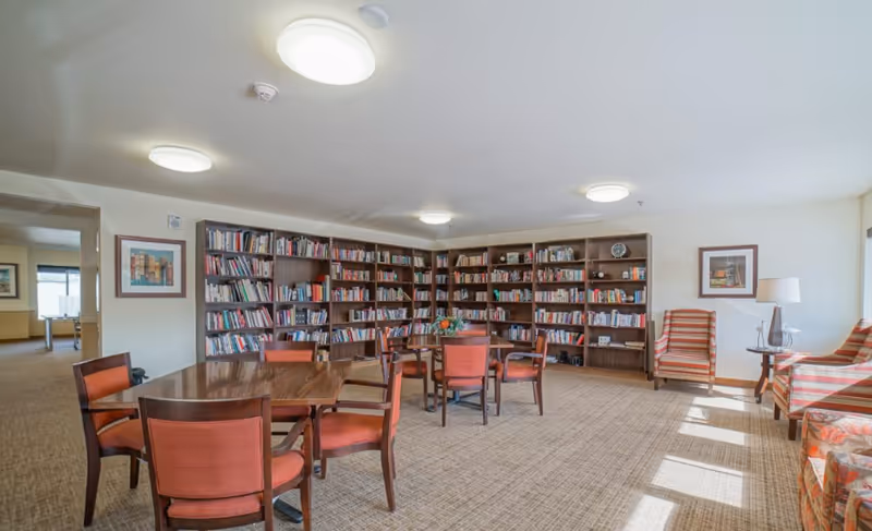 A well-lit room with multiple wooden bookshelves filled with books along the back wall. In the foreground, there are wooden tables with red cushioned chairs arranged around them. To the right, there are two striped armchairs and a small round table with a lamp. The room has a beige carpet and white walls with framed artwork. Large windows on the right allow natural light to enter the space.