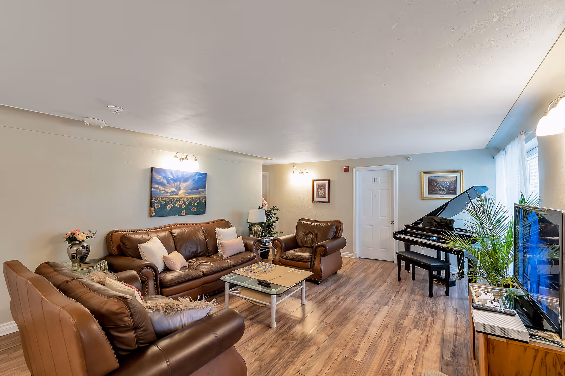 A bright living room with leather sofas and armchairs, a coffee table, a grand piano, television, and potted plants on wood flooring.