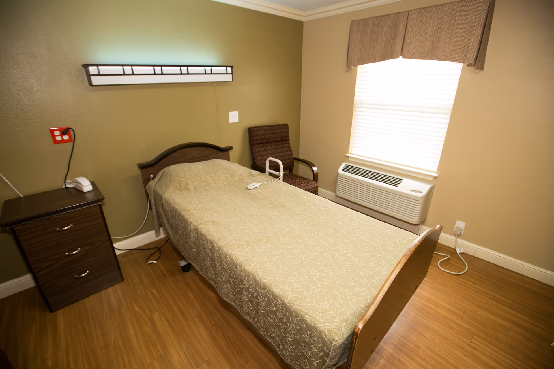A simple bedroom in a healthcare facility with a single bed covered in a beige patterned bedspread, a brown wooden nightstand with a white telephone, a brown cushioned chair, a window with blinds and a beige valance, and a wall-mounted air conditioning unit below the window.
