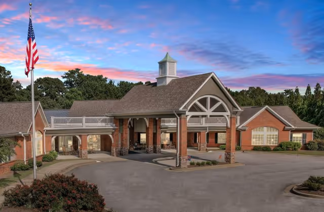 Exterior view of Gaines Park Senior Living facility at sunset, showing a large brick building with a covered entrance, an American flag on a flagpole, and surrounding greenery.
