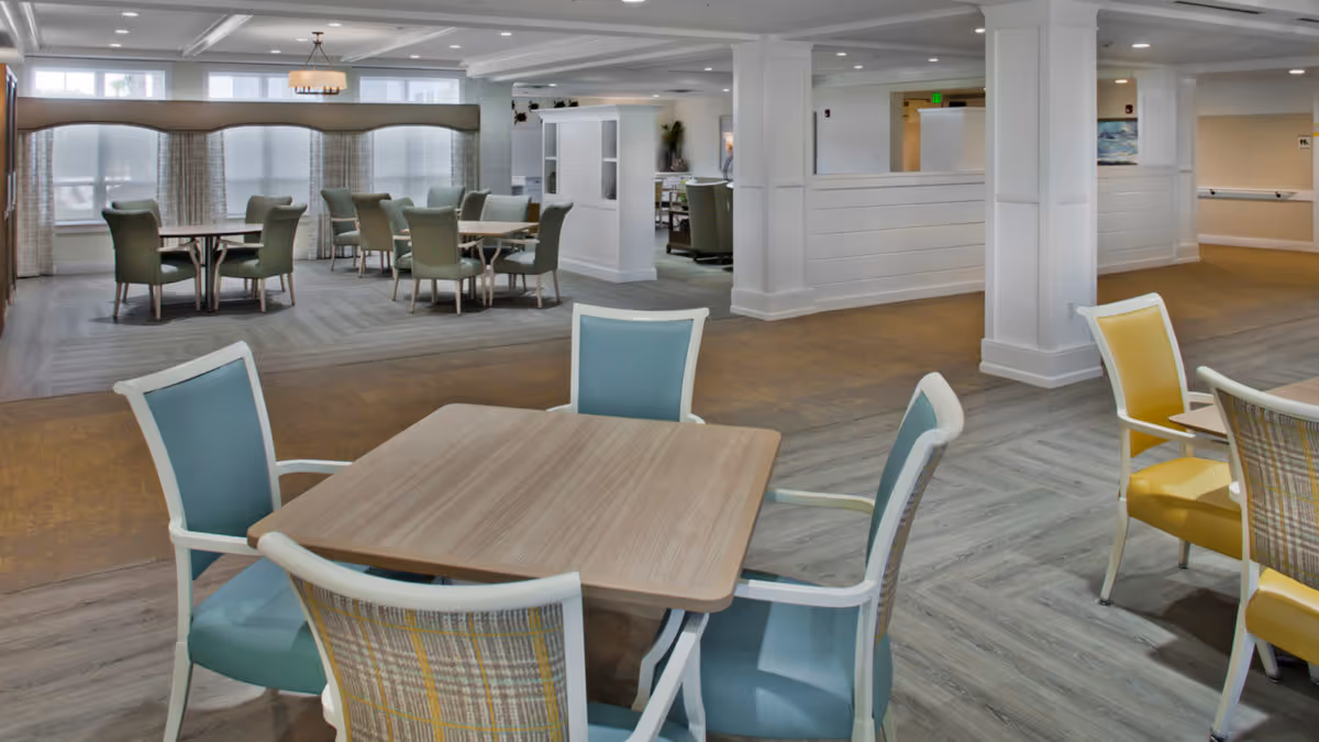 A spacious and well-lit dining area in a senior living facility with multiple tables and chairs arranged neatly. The room features large windows with curtains, light-colored walls, and a combination of wood and carpet flooring. The furniture includes chairs with blue and yellow upholstery and a wooden table in the foreground.