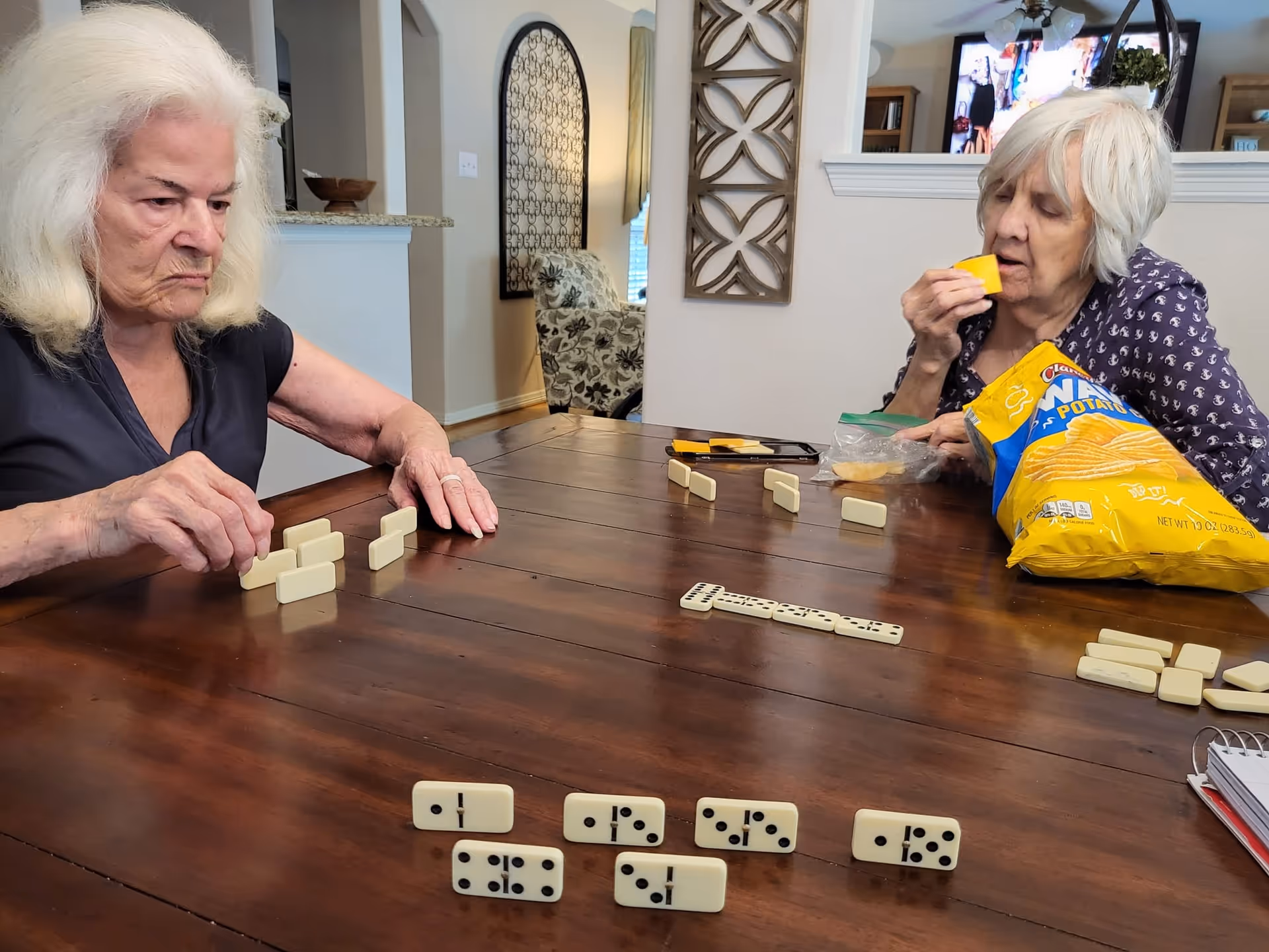 Two elderly women sitting at a wooden table playing dominoes. One woman is arranging her domino pieces while the other is eating a piece of cheese with a bag of Lay's Classic potato chips on the table. The background shows a living area with decorative wall art and a television.