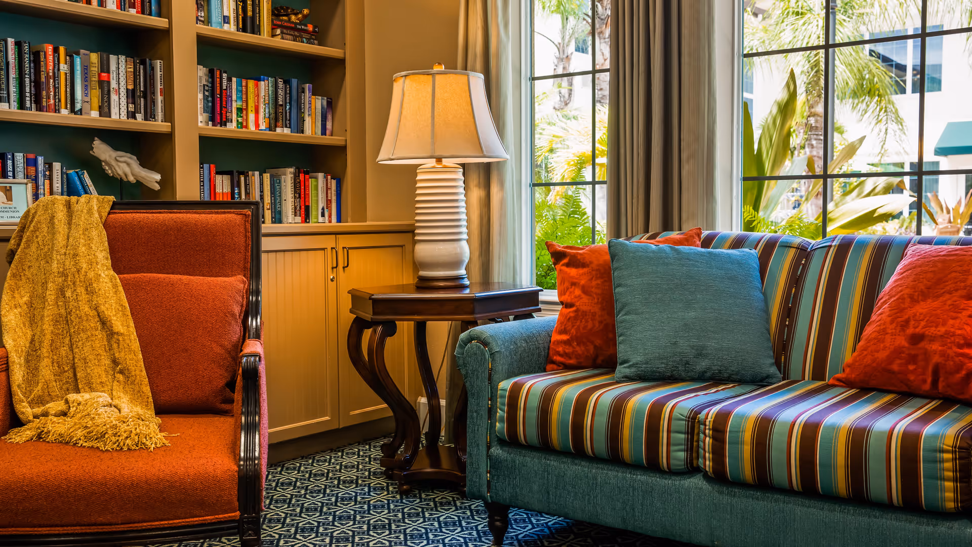 Cozy living room area with a striped sofa adorned with red and blue cushions, an orange armchair with a yellow throw blanket, a wooden side table with a white lamp, and built-in bookshelves filled with books. Large windows let in natural light and show greenery outside.