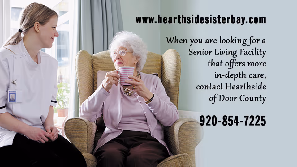 A caregiver in a white uniform is sitting and smiling while talking to an elderly woman with white hair and glasses who is seated in a comfortable armchair holding a striped mug. They are in a well-lit room with a window and curtains in the background.