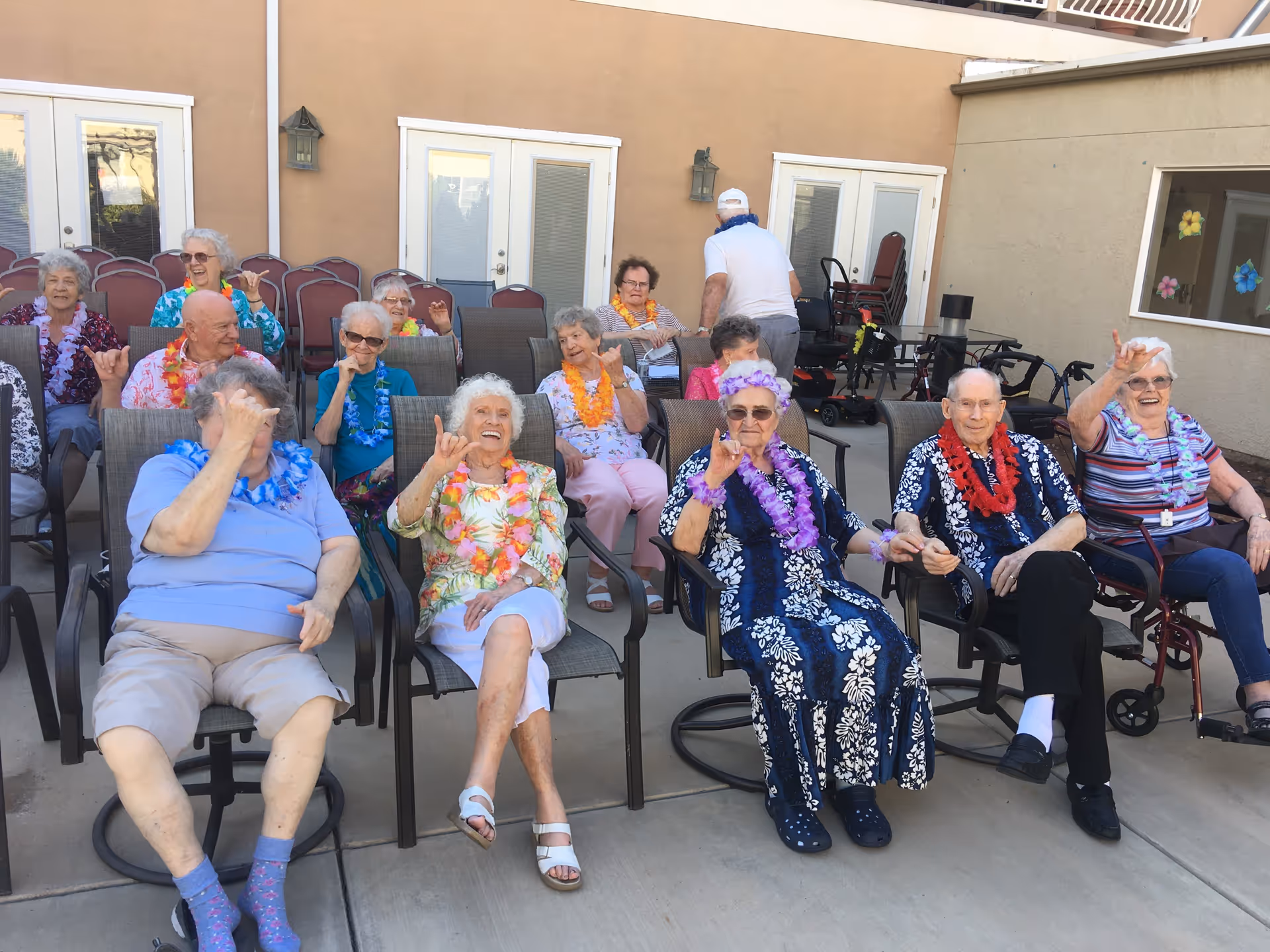 A group of elderly people sitting outdoors on patio chairs, wearing colorful leis and making hand gestures, enjoying a social gathering at an assisted living community.