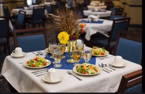 A dining table set for four with plates of salad, glasses of water and wine, white cups and saucers, silverware, and a centerpiece featuring yellow flowers and dried wheat stalks. The background shows more similarly set tables and chairs in a dining room.