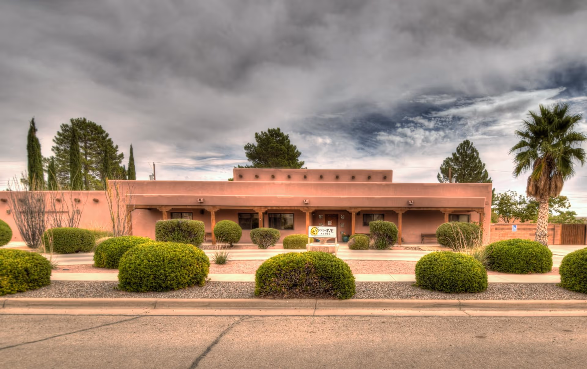 Single-story adobe-style building with desert landscaping, trimmed bushes and a BeeHive Homes sign in front.