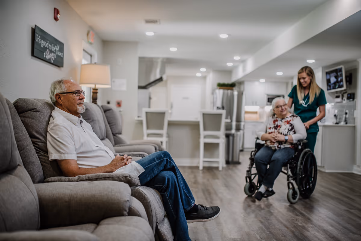 An elderly man sits on a gray recliner sofa in a living room area, while a woman in a wheelchair is being pushed by a caregiver in green scrubs. The background shows a modern kitchen with white cabinets, a refrigerator, and bar stools. The room has wooden flooring and recessed lighting.