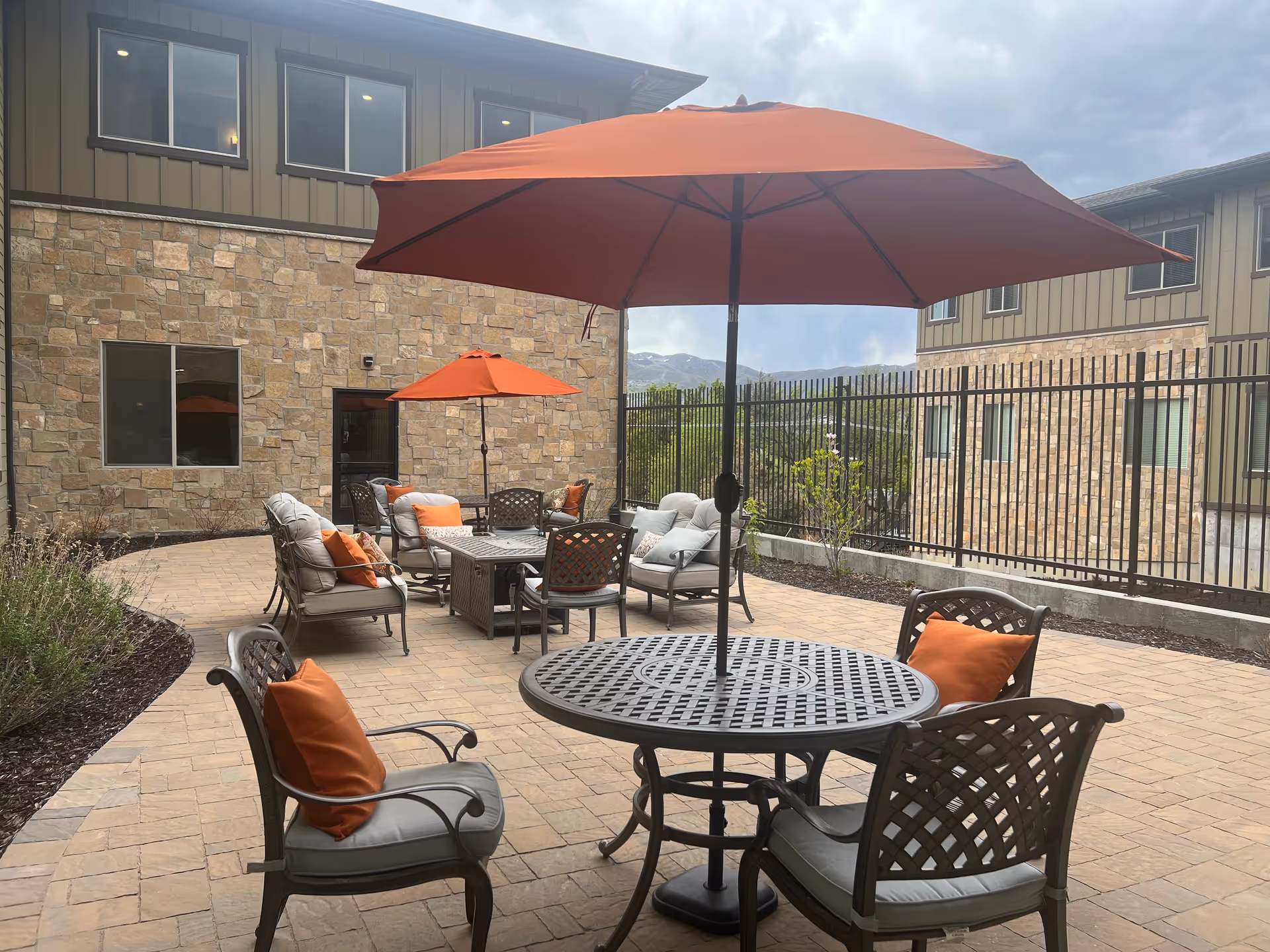 Outdoor patio area with metal tables and cushioned chairs featuring orange pillows. Large orange umbrellas provide shade. The patio is surrounded by a stone building and a black metal fence, with mountains visible in the background under a cloudy sky.
