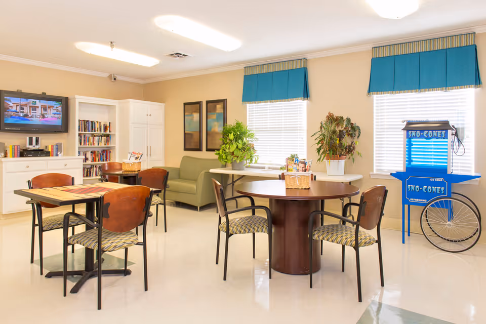 A bright and clean common room with tables and chairs arranged for socializing or activities. There is a green couch against the wall, two windows with blue valances, a blue sno-cones machine, a bookshelf filled with books, a TV mounted on the wall, and some plants and framed artwork decorating the space.
