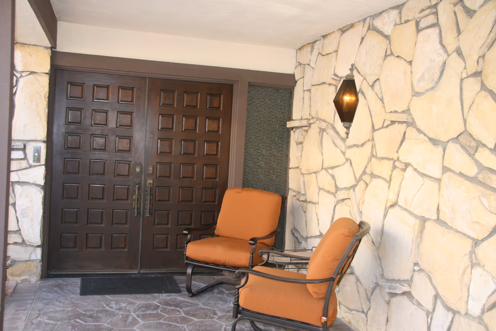 Covered stone entryway with double carved wooden doors, two orange cushioned chairs, and a wall lantern.