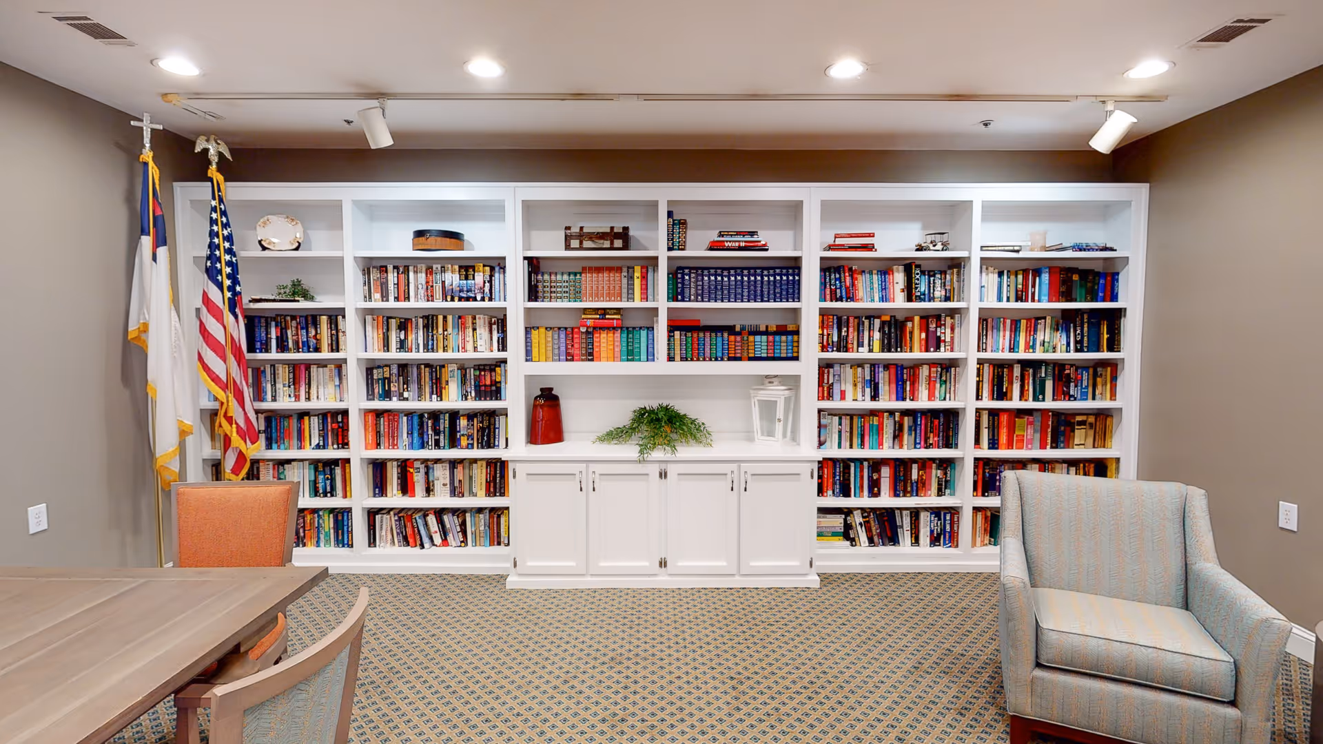 A cozy interior room featuring a large white built-in bookshelf filled with books and decorative items. To the left, two flags stand in the corner. There is a wooden table with chairs on the left side and a cushioned armchair on the right. The room has recessed ceiling lights and a patterned carpet.