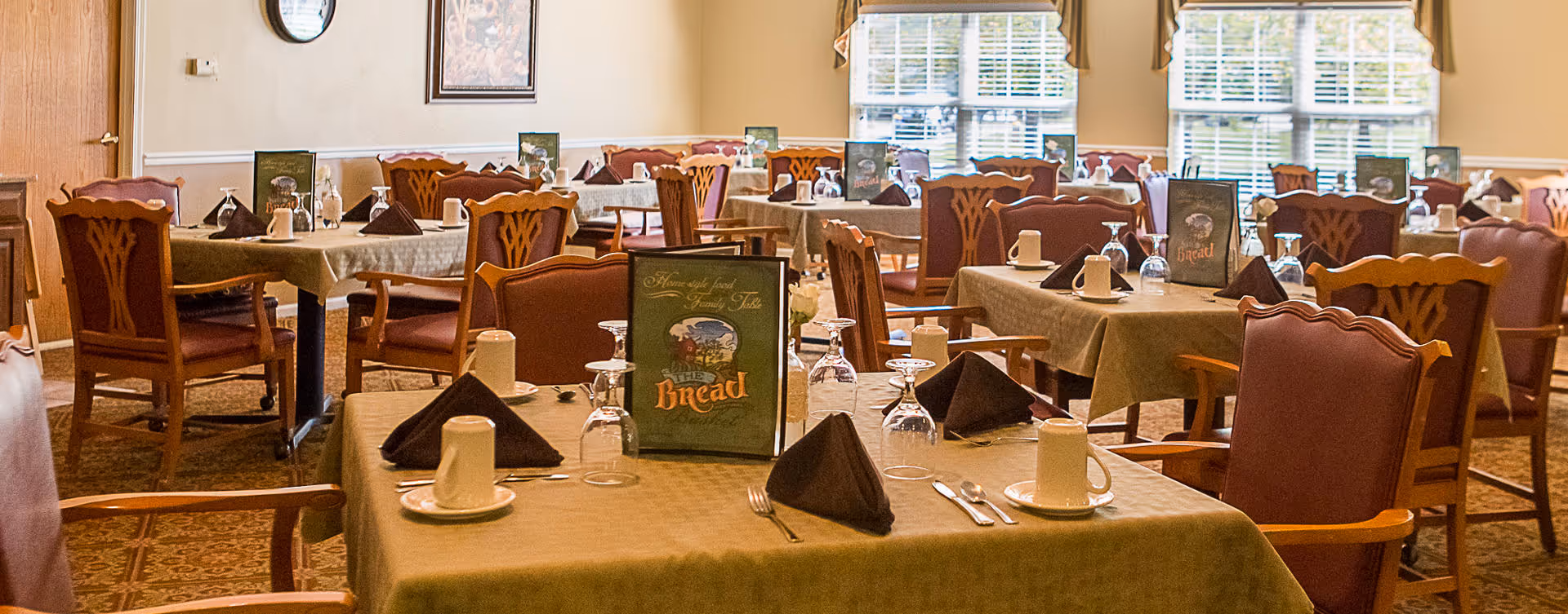 A dining room with multiple tables covered in beige tablecloths, each set with upside-down white cups on saucers, silverware, folded brown napkins, and upside-down wine glasses. The chairs have wooden frames with cushioned seats and backs. There are large windows with blinds and valances letting in natural light, and a framed picture on the wall.