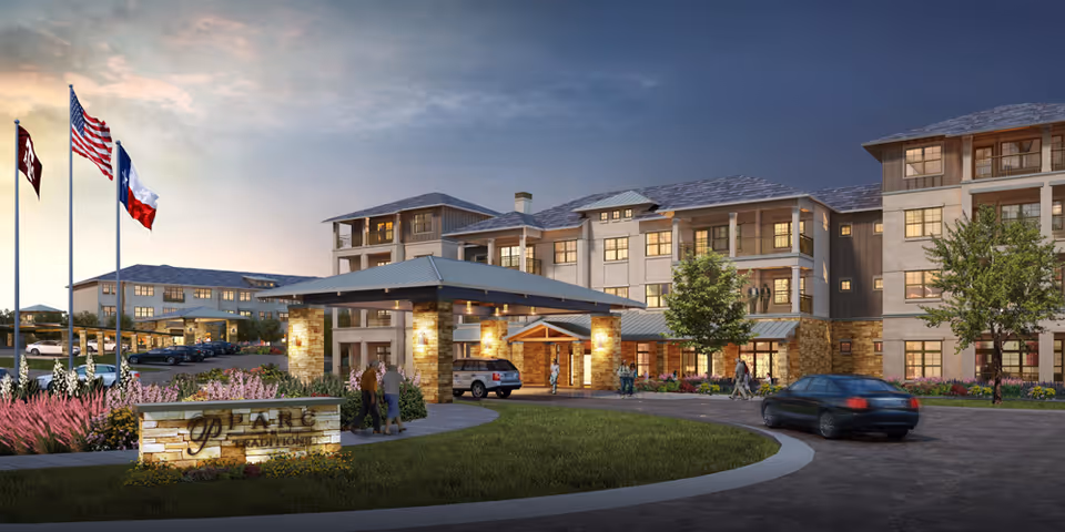 Exterior front entrance of a multi-story senior living community at dusk with flags, a landscaped roundabout, vehicles and people.
