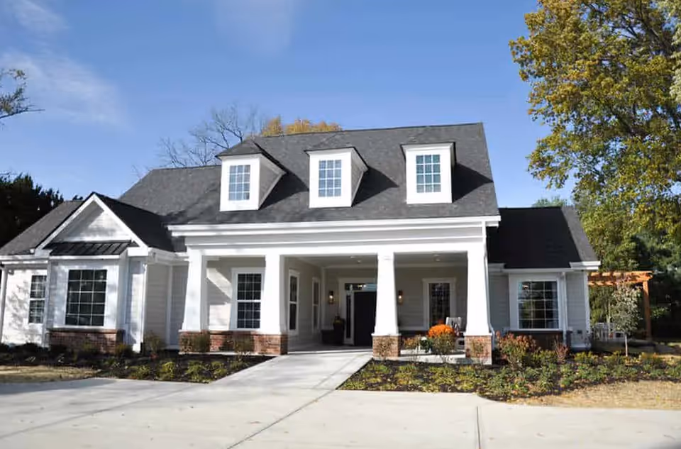 Front exterior view of a white single-story building with a dark gray roof, three dormer windows, a covered entrance supported by four white columns, and landscaped garden beds with shrubs and flowers under a clear blue sky.
