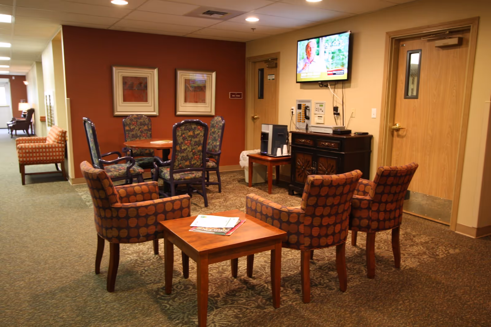 A cozy seating area in a senior living facility with patterned armchairs arranged around a wooden table with magazines on it. In the background, there is a round table with floral upholstered chairs against a red accent wall with two framed pictures. A TV is mounted on the wall above a cabinet with a coffee machine and disposable cups. Two wooden doors are visible on the right side.