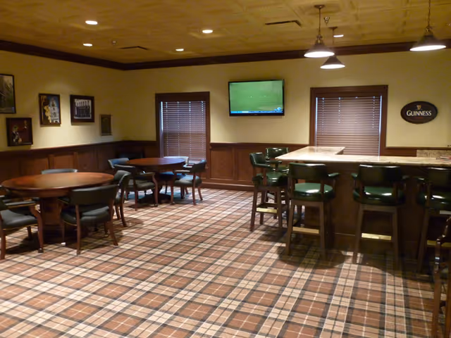 Interior view of a lounge area with plaid carpet, wooden round tables with chairs, a bar counter with green cushioned bar stools, two windows with blinds, a wall-mounted TV, and framed pictures on the wall.