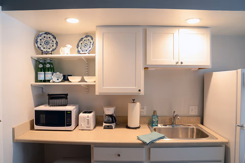Small kitchenette with white cabinets, a microwave, toaster, coffee maker, sink and decorative plates on open shelves.