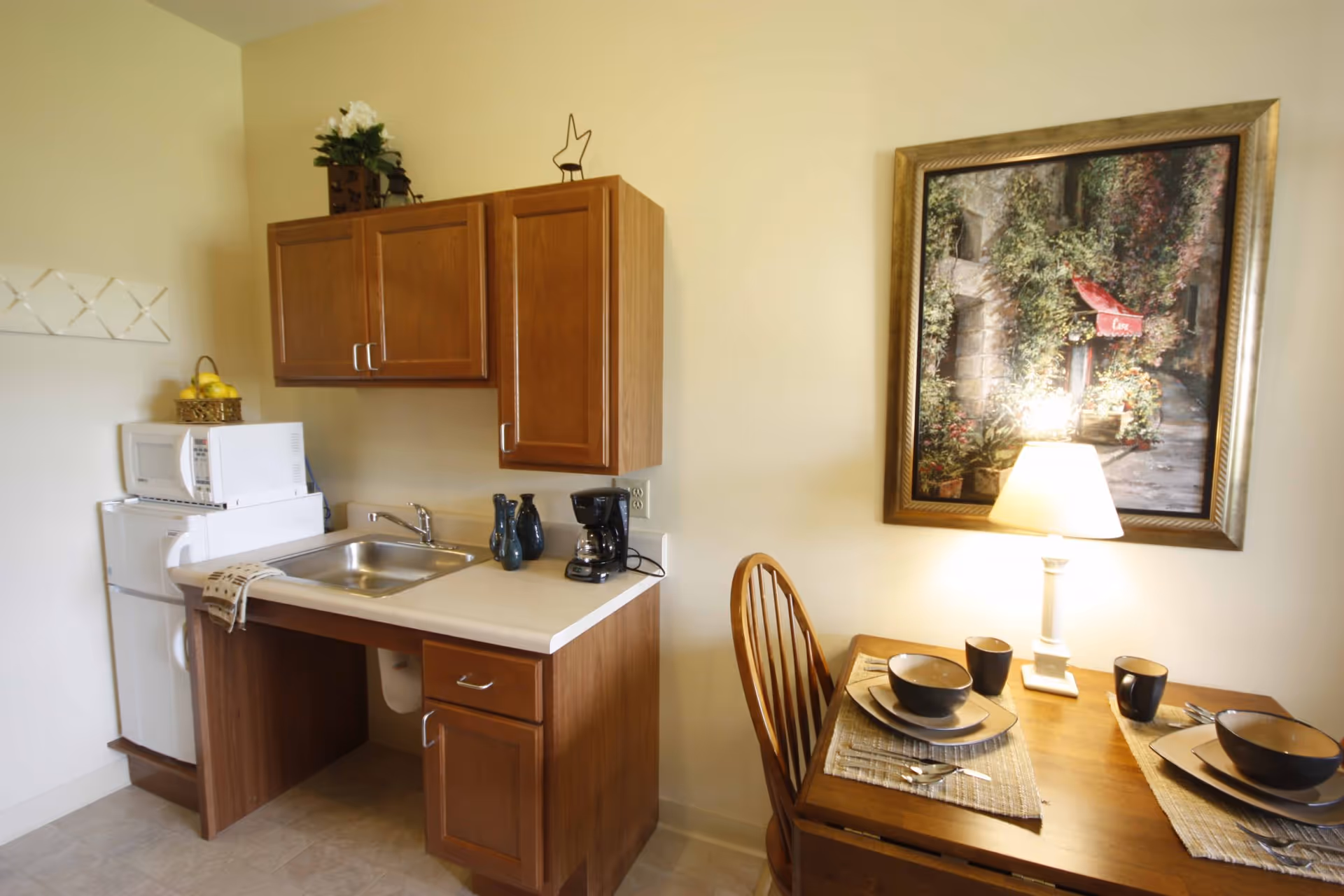 Small kitchenette area with wooden cabinets, a sink, a coffee maker, a microwave, and a mini refrigerator. Next to the kitchenette is a wooden dining table set for two with bowls, plates, cups, and silverware. A table lamp is on the dining table, and a framed painting of a garden scene hangs on the wall above it.