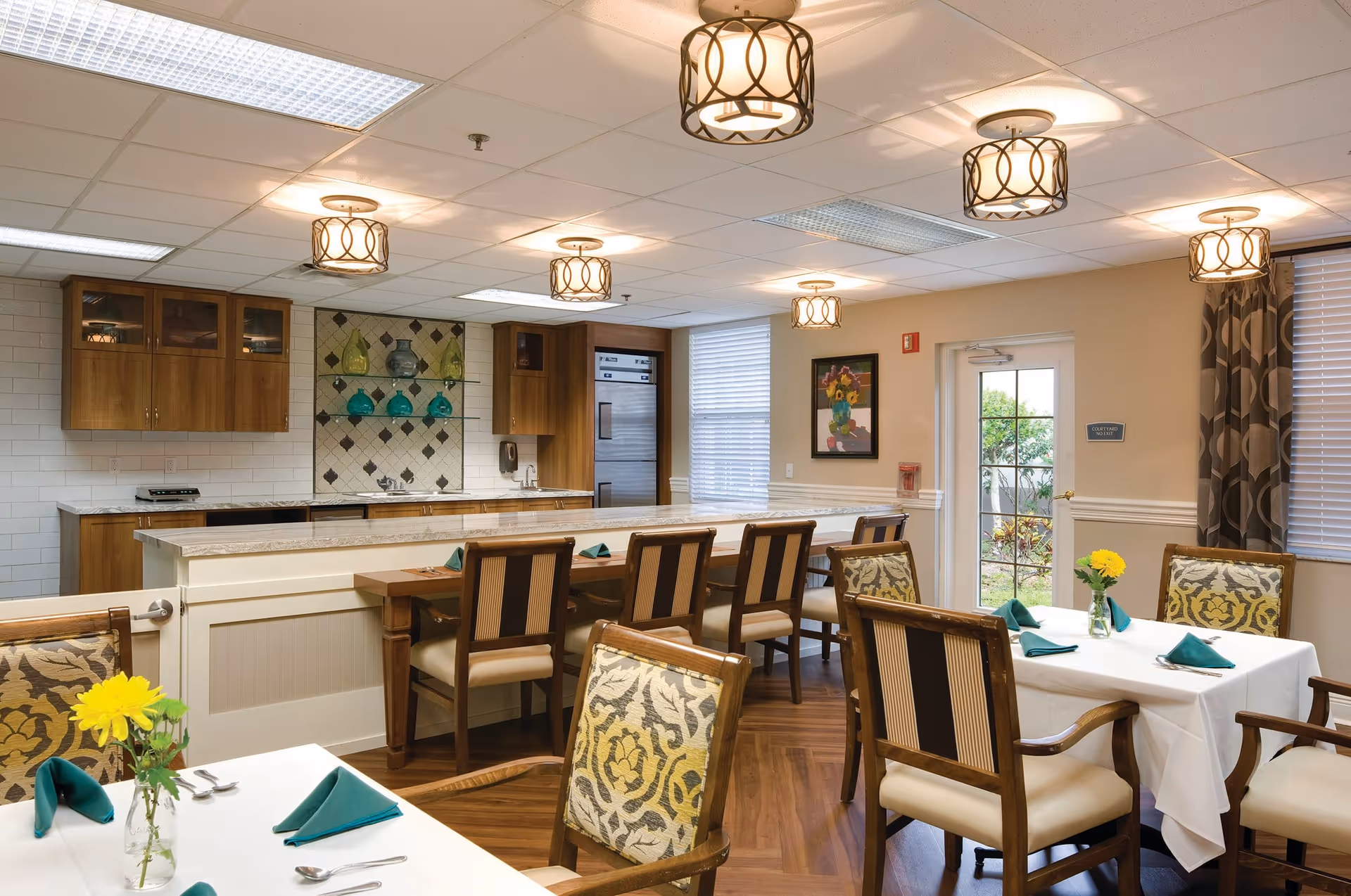 A well-lit dining area with wooden chairs featuring patterned upholstery around tables covered with white tablecloths and green napkins. The room includes a kitchen counter with bar stools, wooden cabinets, decorative glass vases on shelves, and ceiling lights with circular metal designs. A door with glass panels leads outside, and a framed floral painting hangs on the wall.
