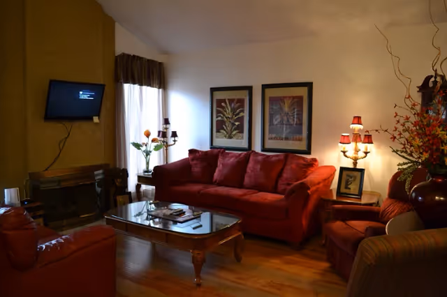 A cozy living room with a red sofa and matching armchairs, a glass coffee table with books and magazines, a wall-mounted TV, a fireplace, two framed botanical prints on the wall, a window with curtains, and decorative lamps and flowers.