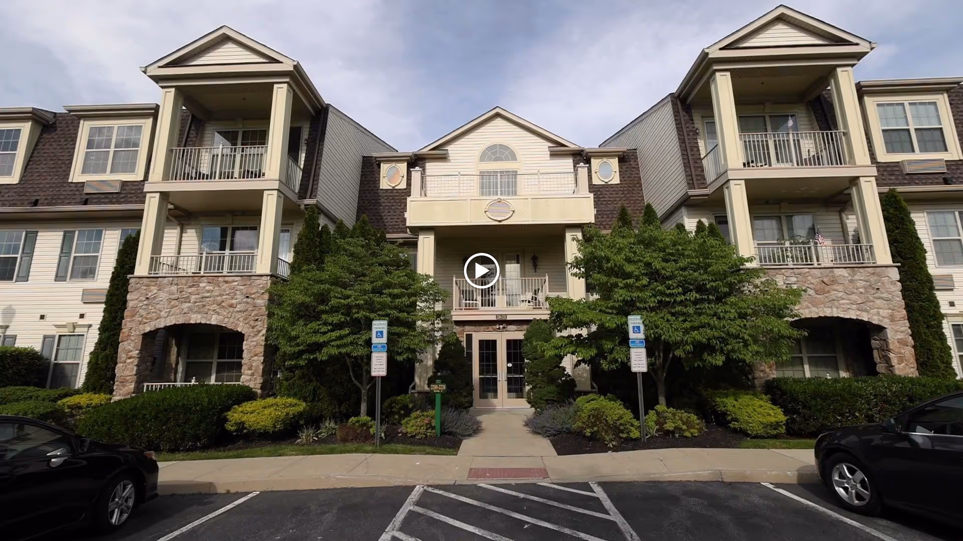Front exterior view of a multi-story senior living facility building with balconies, stone and siding facade, surrounded by landscaped greenery and two handicap parking signs in front.