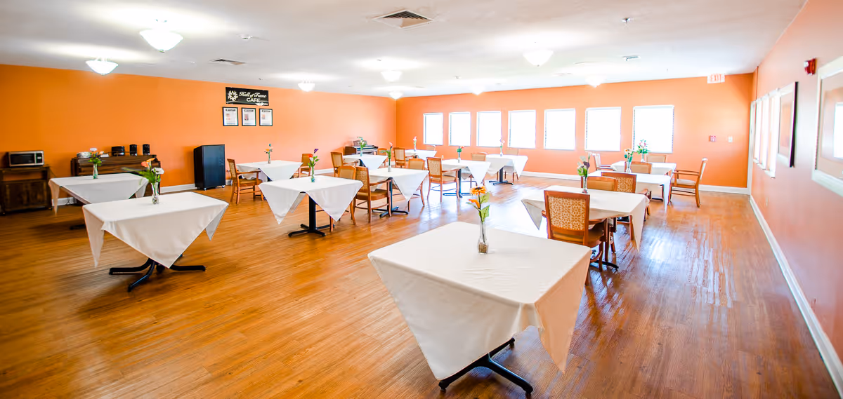 A bright dining room with several tables covered in white tablecloths, each with a small vase of flowers. The room has orange walls, wooden flooring, multiple windows letting in natural light, and a sign on the far wall that reads 'Staff & Visitors Cafe'.