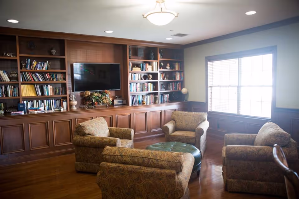 A cozy living room area with four patterned armchairs arranged around a green ottoman. The room features wooden built-in bookshelves filled with books and decorative items, a flat-screen TV mounted on the wall, and a large window letting in natural light.