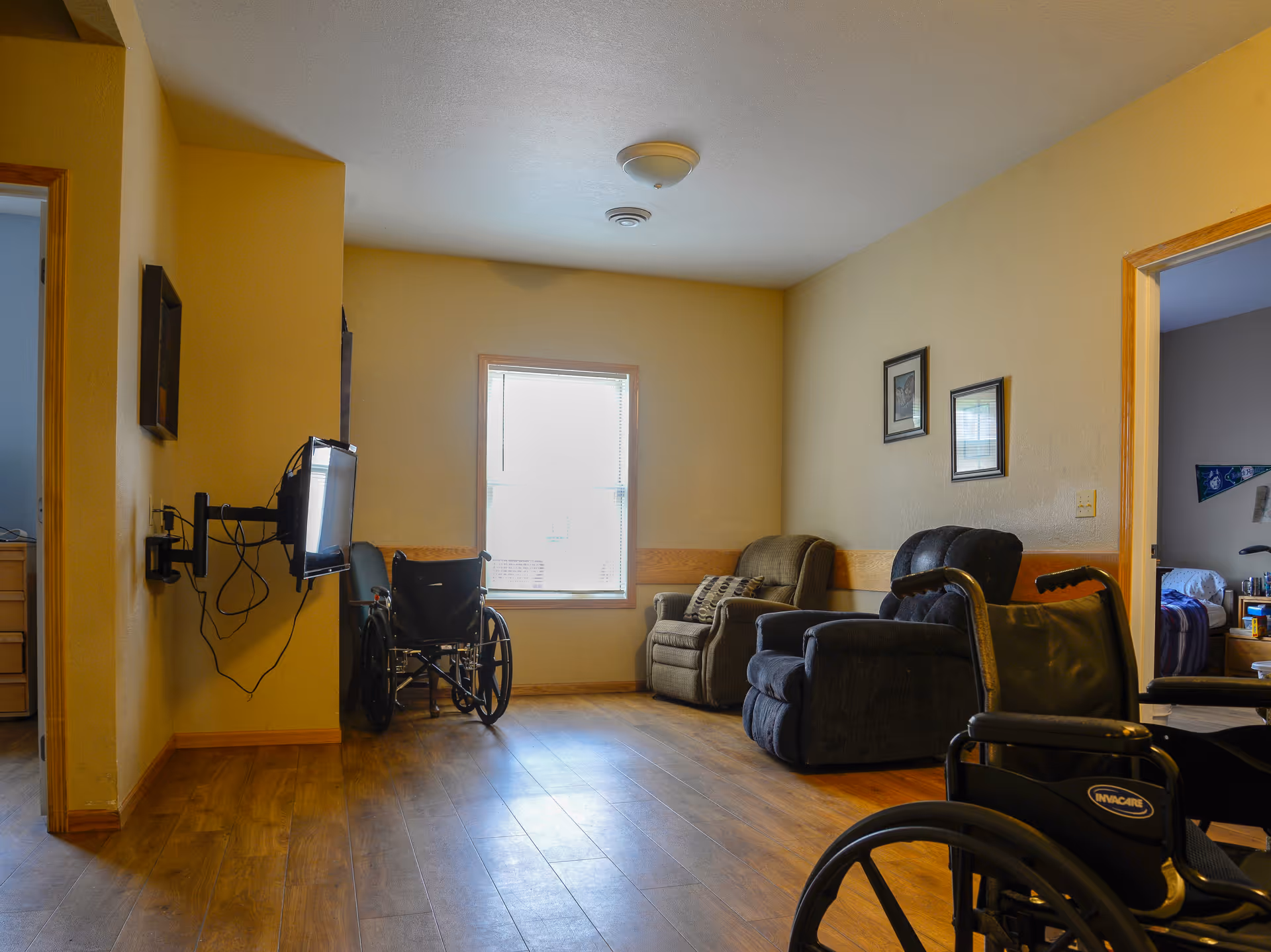 A small living room area in a senior living facility with two wheelchairs, two armchairs, a wall-mounted TV, and a window letting in natural light. The room has wooden flooring and beige walls with two framed pictures. Adjacent rooms are partially visible.