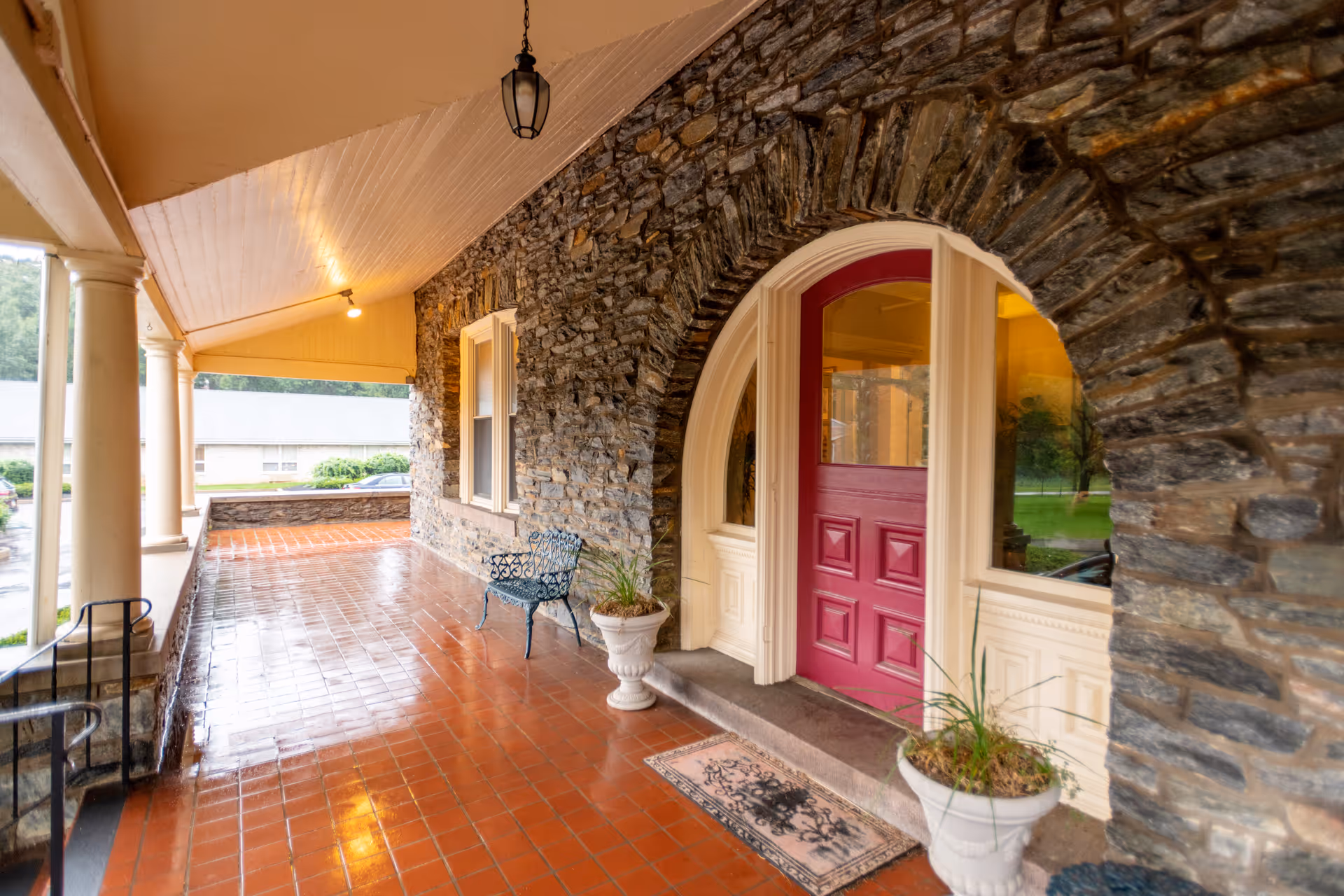 Covered porch area with a red door set in a stone wall, two white planters with plants, a black metal bench, and a tiled floor. The porch has white columns and a ceiling light fixture.
