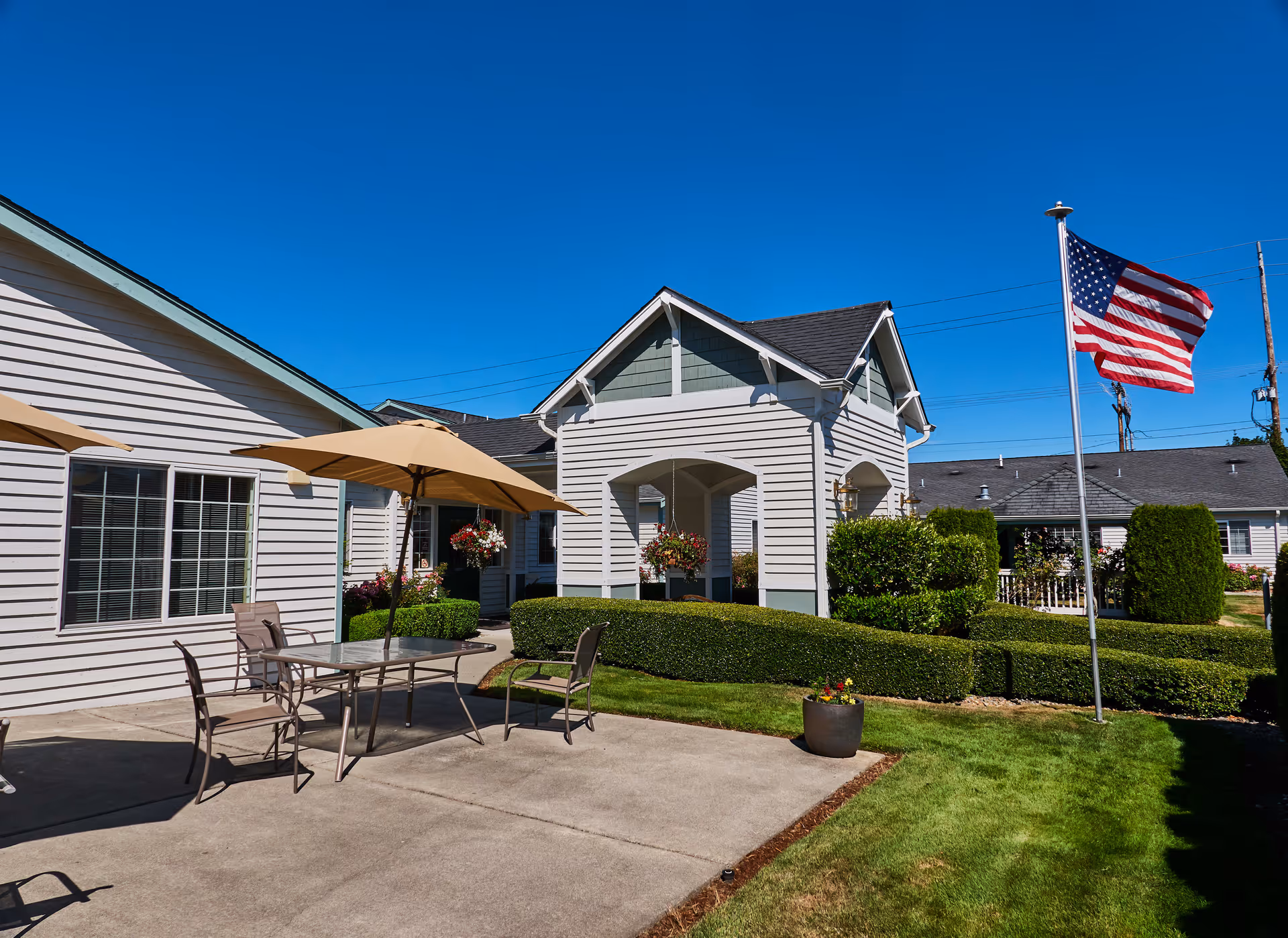 Outdoor patio area at South Pointe Assisted Living with a table, chairs, and umbrellas on a concrete surface. The building exterior is light-colored with green trim, surrounded by neatly trimmed bushes and a well-maintained lawn. An American flag is flying on a flagpole under a clear blue sky.