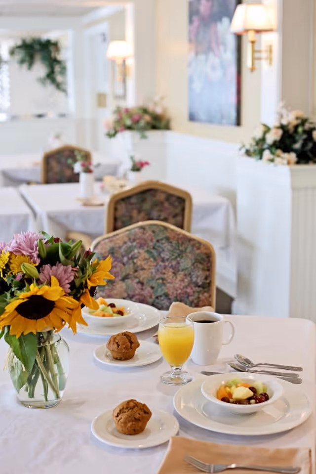 A dining table set for breakfast with two plates of fruit salad, two muffins on small plates, a glass of orange juice, and a cup of coffee. A vase with a bouquet of sunflowers and other flowers is on the table. The background shows more tables and chairs with floral upholstery in a bright, softly lit dining room.