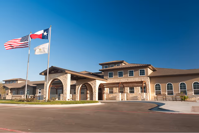 Exterior view of Stoney Brook of Hewitt facility under a clear blue sky, featuring a stone and stucco building with arched entryways, three flagpoles displaying the American flag, Texas state flag, and a white flag, and a curved driveway in front.