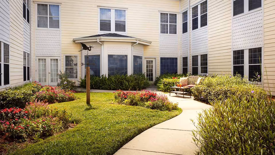 A sunny outdoor courtyard area at Colliers Assisted Living at Port Orange featuring a curved concrete pathway surrounded by green grass, colorful flower beds, and shrubs. There is a wooden post with a bird-shaped weather vane in the center, and a cushioned bench with a small table holding drinks is placed along the pathway near the building. The building exterior is light yellow with multiple windows.