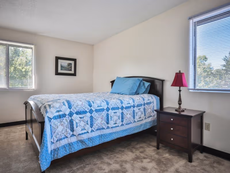A bedroom with a bed covered in a blue and white patterned quilt and two blue pillows. There is a dark wooden nightstand with a red lamp on it next to the bed. Two windows with blinds let in natural light, and a framed picture hangs on the wall above the bed.